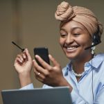 Mulher usando headset enquanto consulta um smartphone e trabalha em um laptop em um ambiente de escritório.