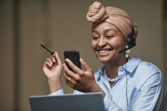 Mulher usando headset enquanto consulta um smartphone e trabalha em um laptop em um ambiente de escritório.