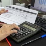 Pessoa calcula dados financeiros com uma calculadora e documentos sobre uma mesa de escritório.
