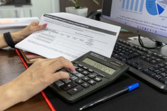 Pessoa calcula dados financeiros com uma calculadora e documentos sobre uma mesa de escritório.