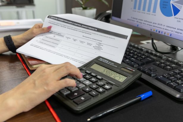 Pessoa calcula dados financeiros com uma calculadora e documentos sobre uma mesa de escritório.