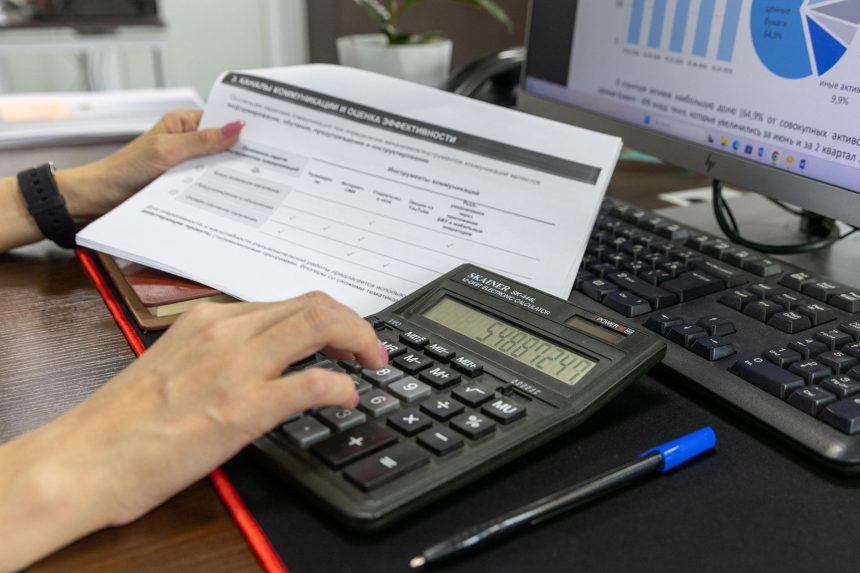 Pessoa calcula dados financeiros com uma calculadora e documentos sobre uma mesa de escritório.